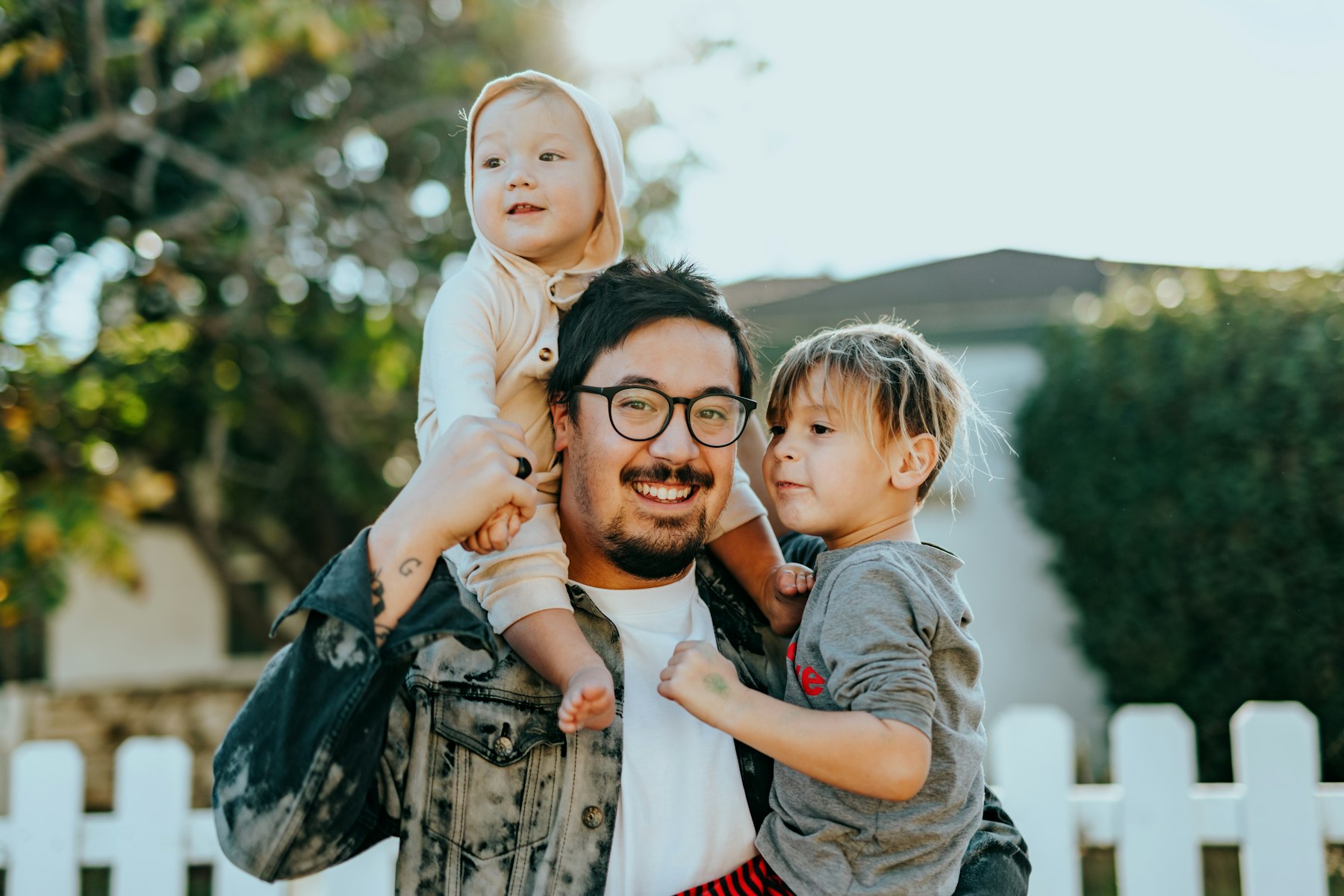 A smiling father outdoors carrying one child on his shoulders and holding another